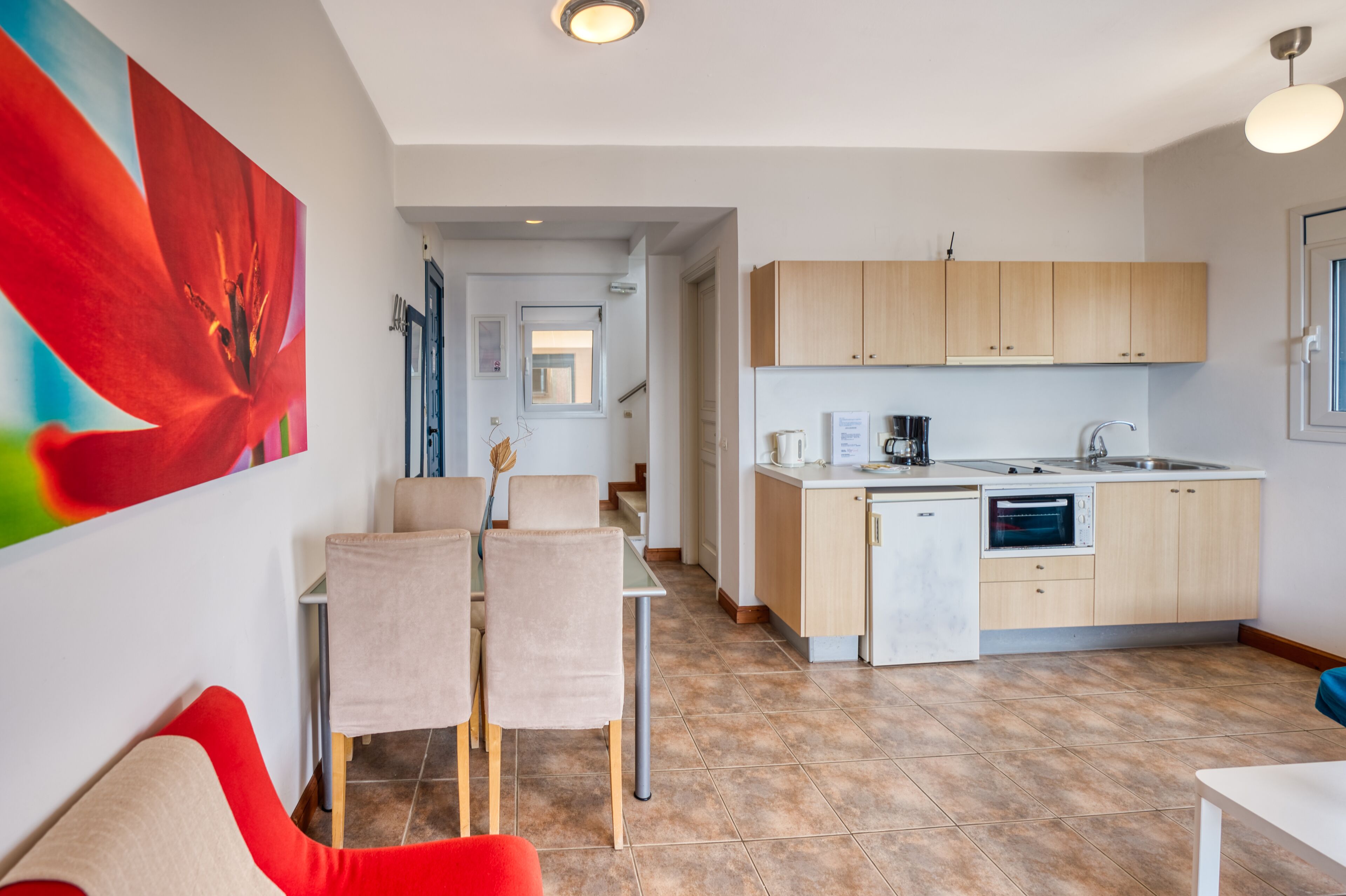 A modern kitchen with light wood cabinets, a built-in oven, and a sink. There is a dining area with a table and chairs, and a large abstract painting on the wall.