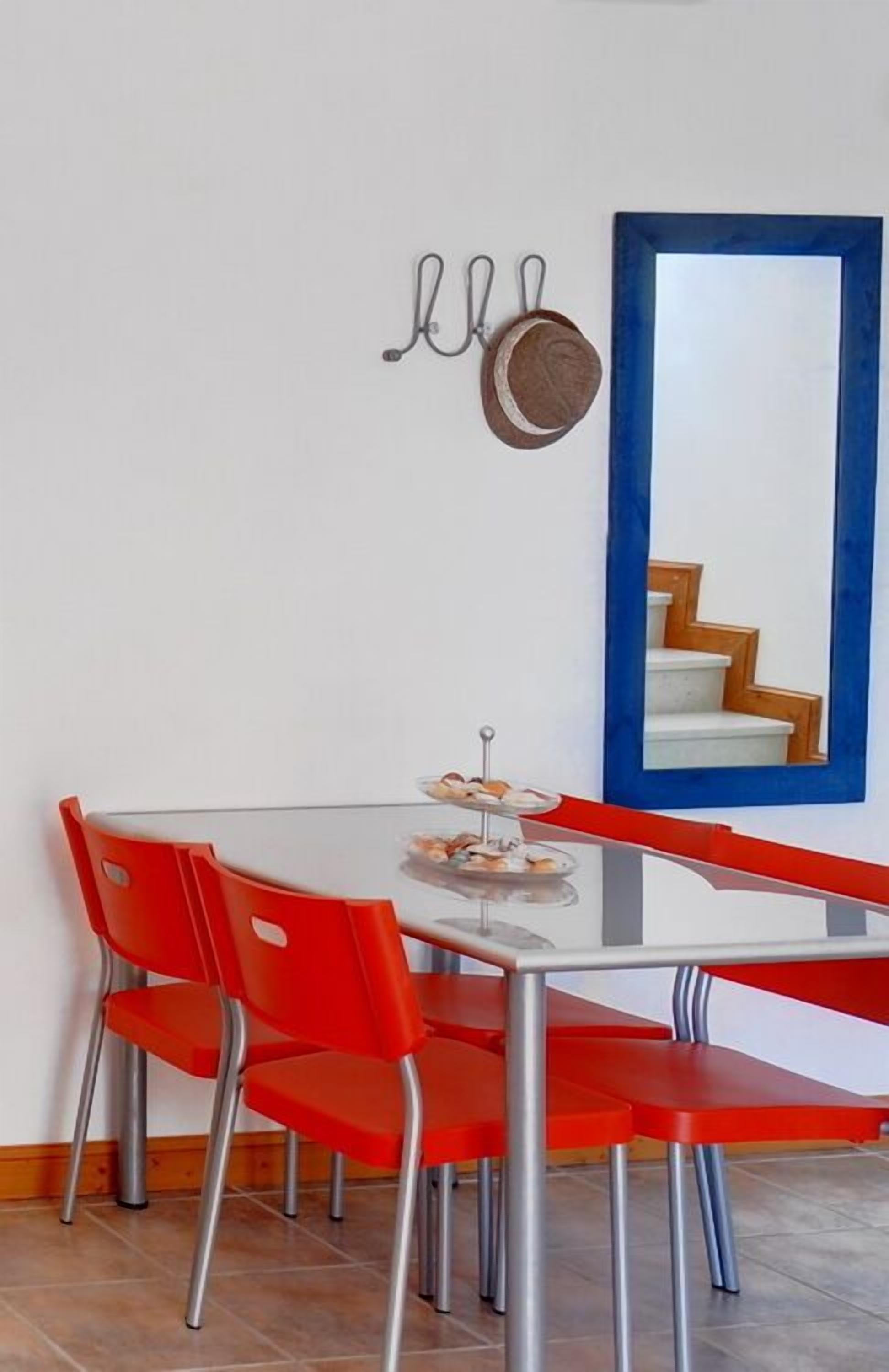A dining area with a glass table, red chairs, a small dessert display, a wall-mounted pot, and a blue-framed mirror reflecting a staircase.