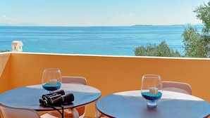 A balcony with two round tables, each with a glass containing blue beads and a candle. The view includes a body of water and distant land.