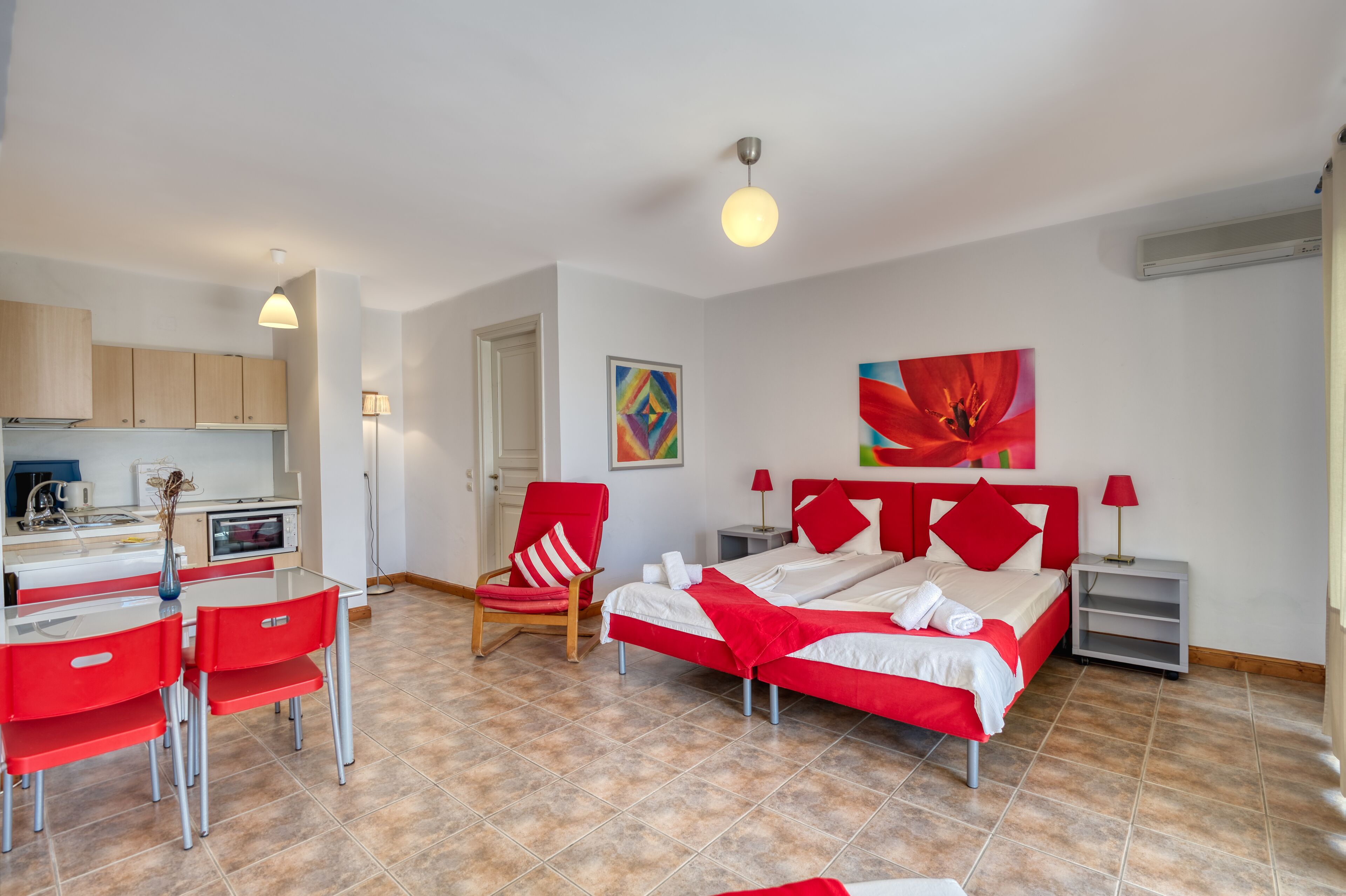 A modern hotel room with a red and white color scheme, a bed, a desk with a chair, a small kitchen area, and a bathroom.