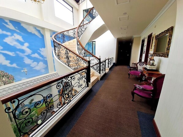 Una imponente escalera con barandales de metal ornamentados, que conduce a un balcón con un mural que representa el cielo y las nubes.