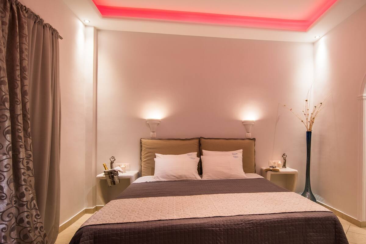 Una cama bien hecha con cabecera, dos mesitas de noche con lámparas, un jarrón con plantas secas y una cortina a la izquierda.