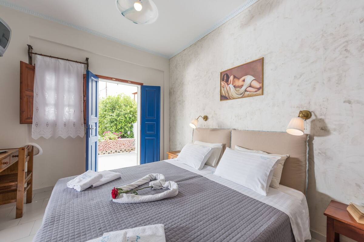 Un dormitorio con cama, almohadas, mesita de noche, lámpara, un cuadro, una ventana con cortinas y vistas a zonas verdes.