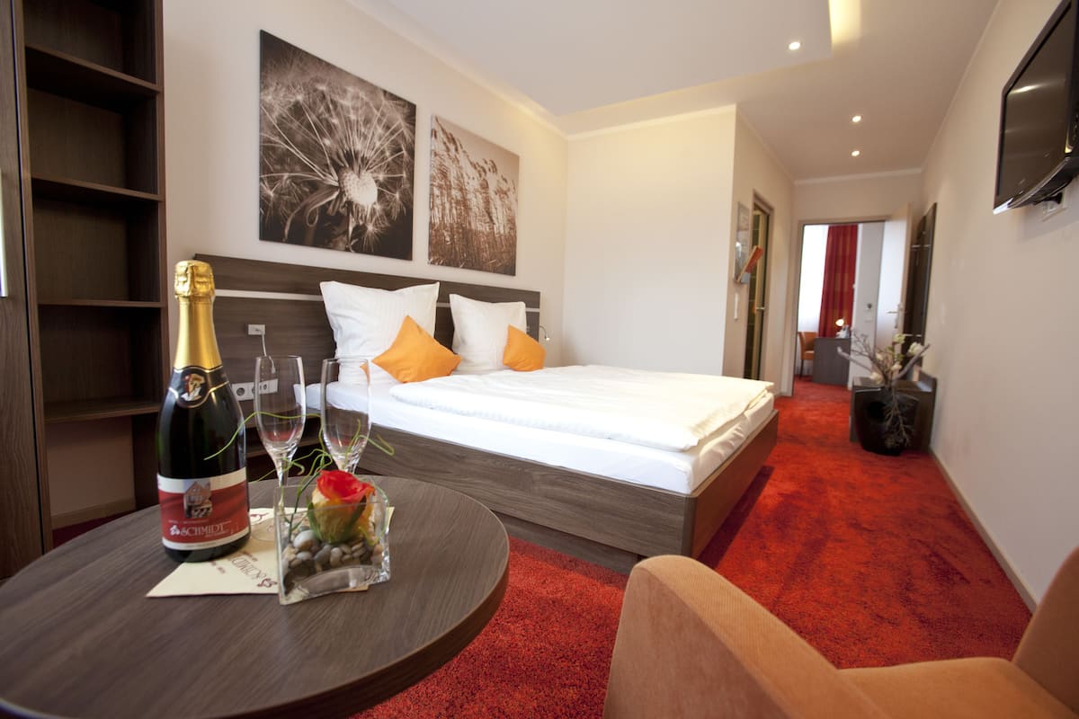 Una habitación de hotel con una cama, una mesita de noche con una botella de champán y un pequeño florero con flores.