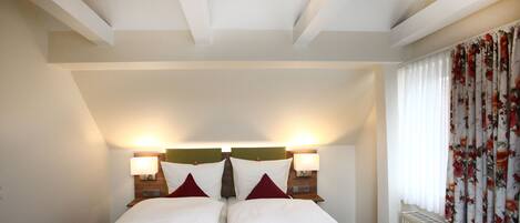 Una habitación de hotel con una cama grande, dos lámparas de noche, un radiador y una ventana con cortinas florales.
