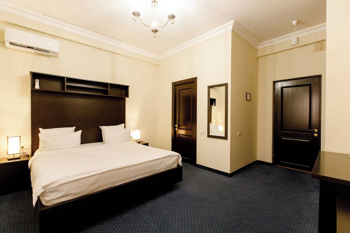 A hotel room with a bed, bedside tables, a mirror, a door, and an air conditioning unit.