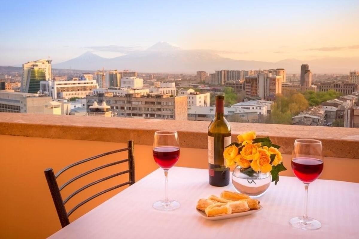A table with two wine glasses, a bottle of wine, and a vase of flowers on a balcony overlooking a cityscape with mountains in the background.