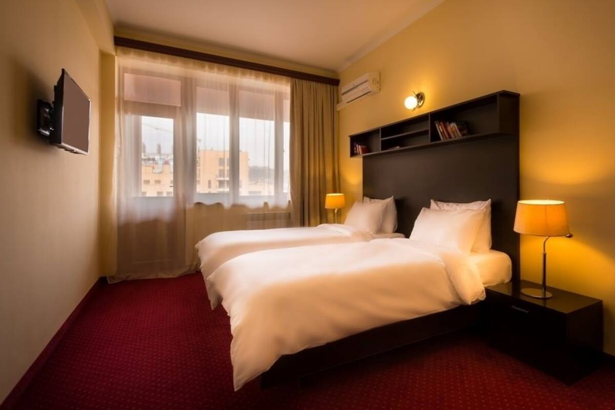 A hotel room with two beds, a TV mounted on the wall, a bookshelf, and a window with curtains.