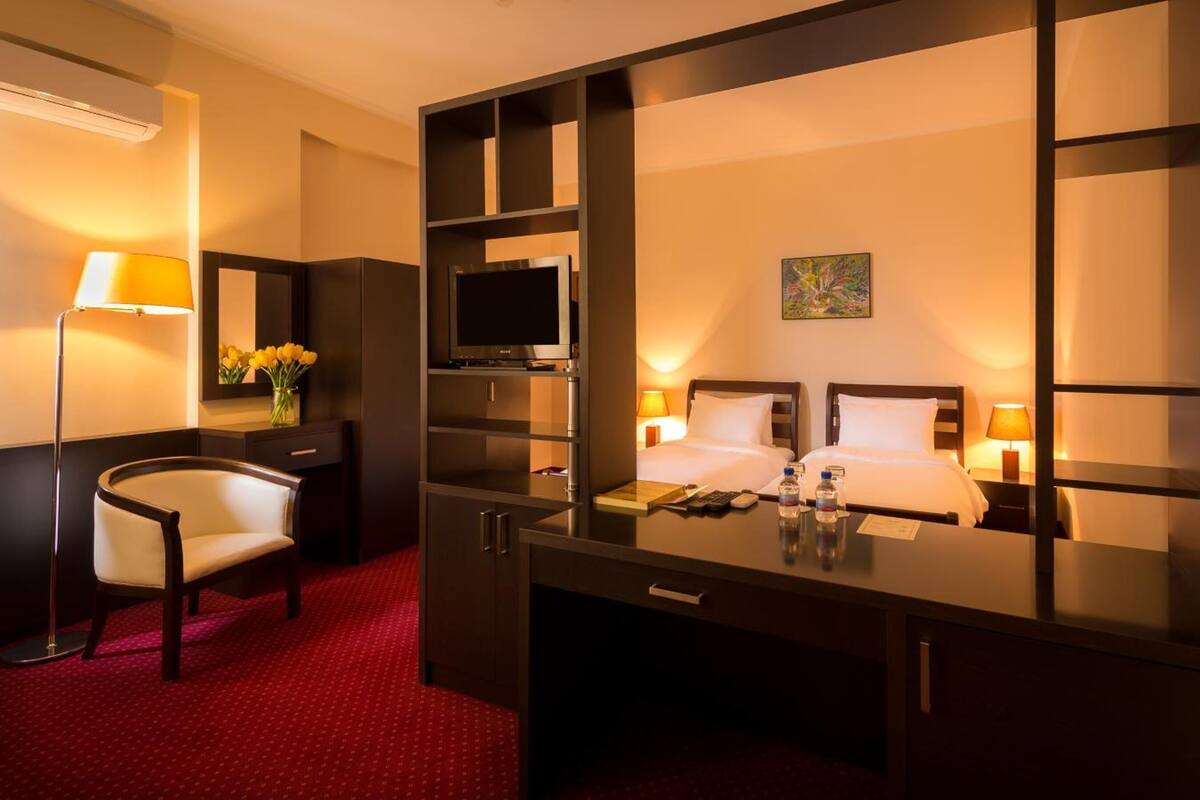 A hotel room with a desk, chair, bed, TV, and artwork on the wall.