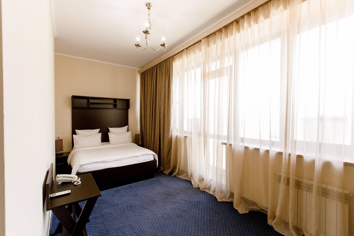 A hotel room with a large bed, a bedside table with a phone, and a window with sheer curtains.