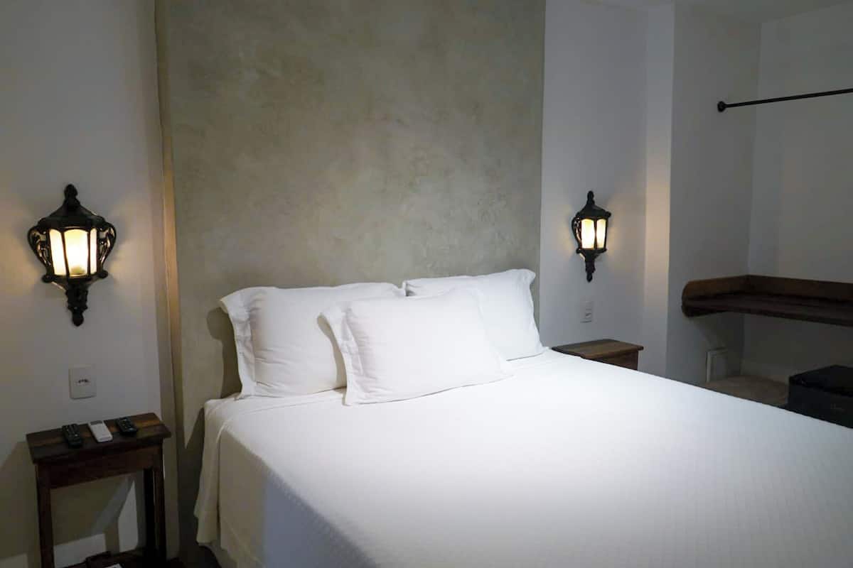 En snyggt bäddad säng med vita sängkläder i ett hotellrum. Det finns två sängbord, var och en med en lampa och en fjärrkontroll på det ena. Väggarna är enfärgade med en strukturerad yta, och det står en träbänk längs väggen.