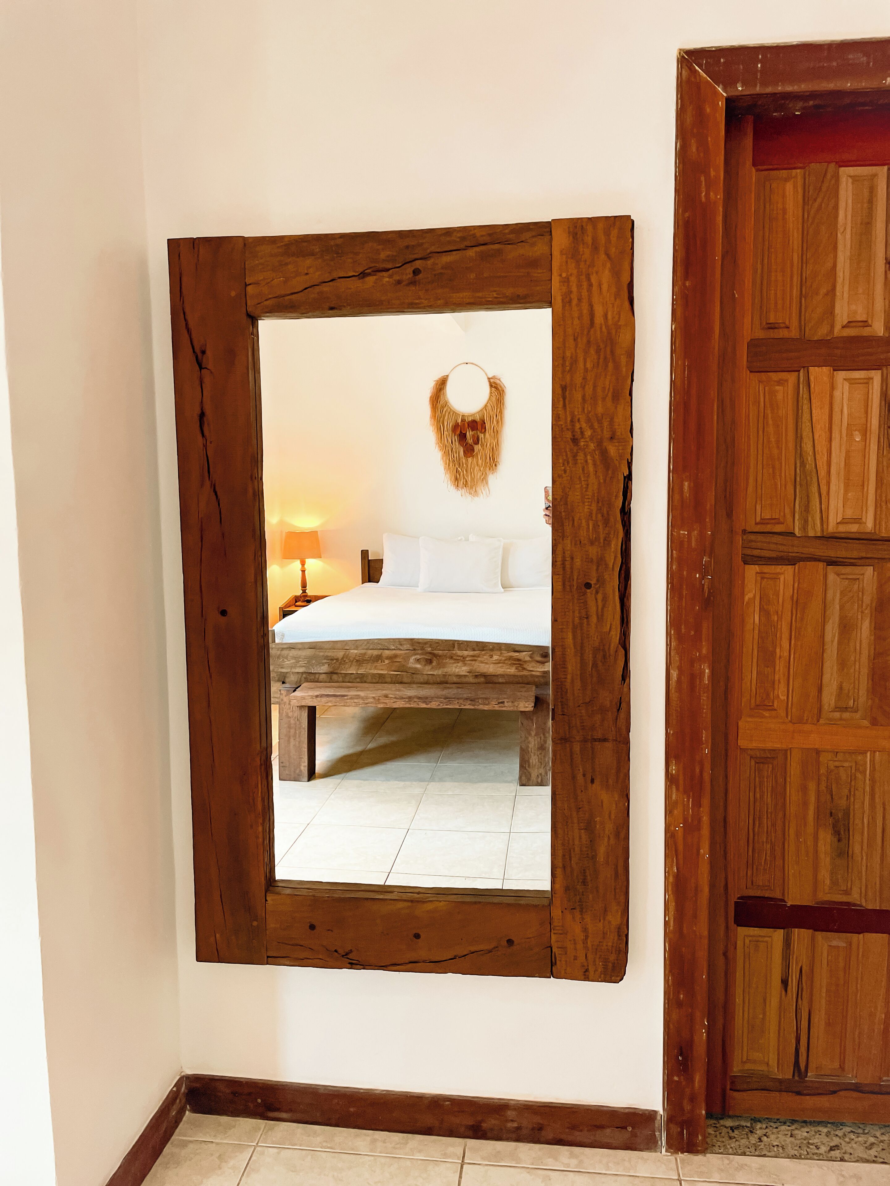 Un espejo de madera rústica refleja una cama, un adorno de pared y una puerta de madera.