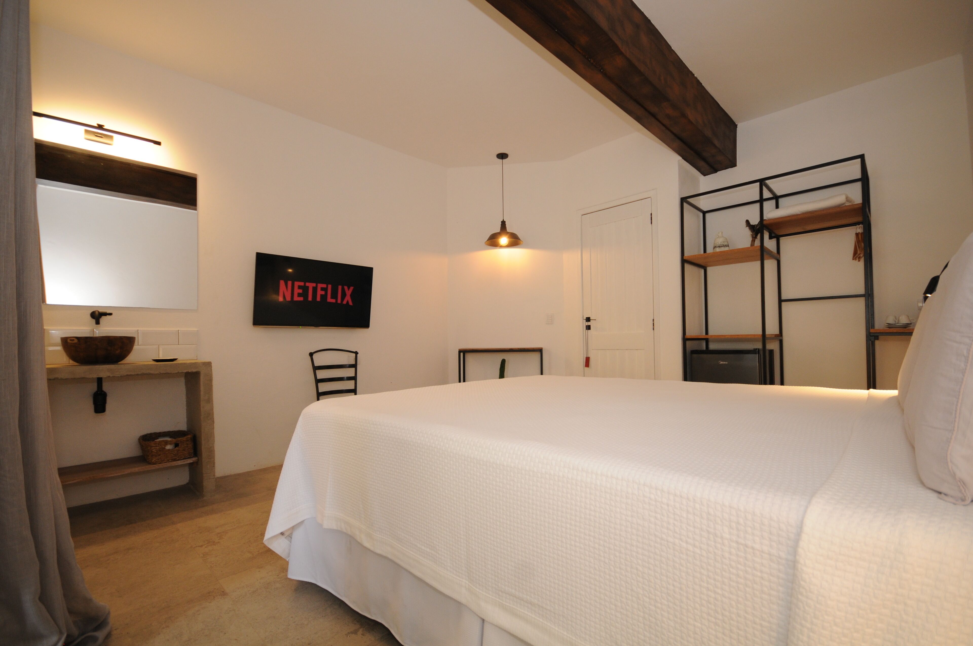 A bedroom with a large bed, a TV mounted on the wall, a wooden shelf, and a hanging light fixture.