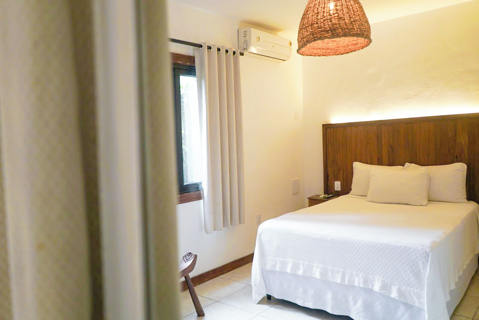 Une chambre d’hôtel avec un grand lit, une tête de lit en bois, une table de chevet, une lampe et une fenêtre avec des rideaux.