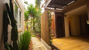 Un étroit sentier mène à une porte en bois, bordée d’un cactus et d’une clôture en bambou, avec une zone dallée de pierres et un plancher en bois.