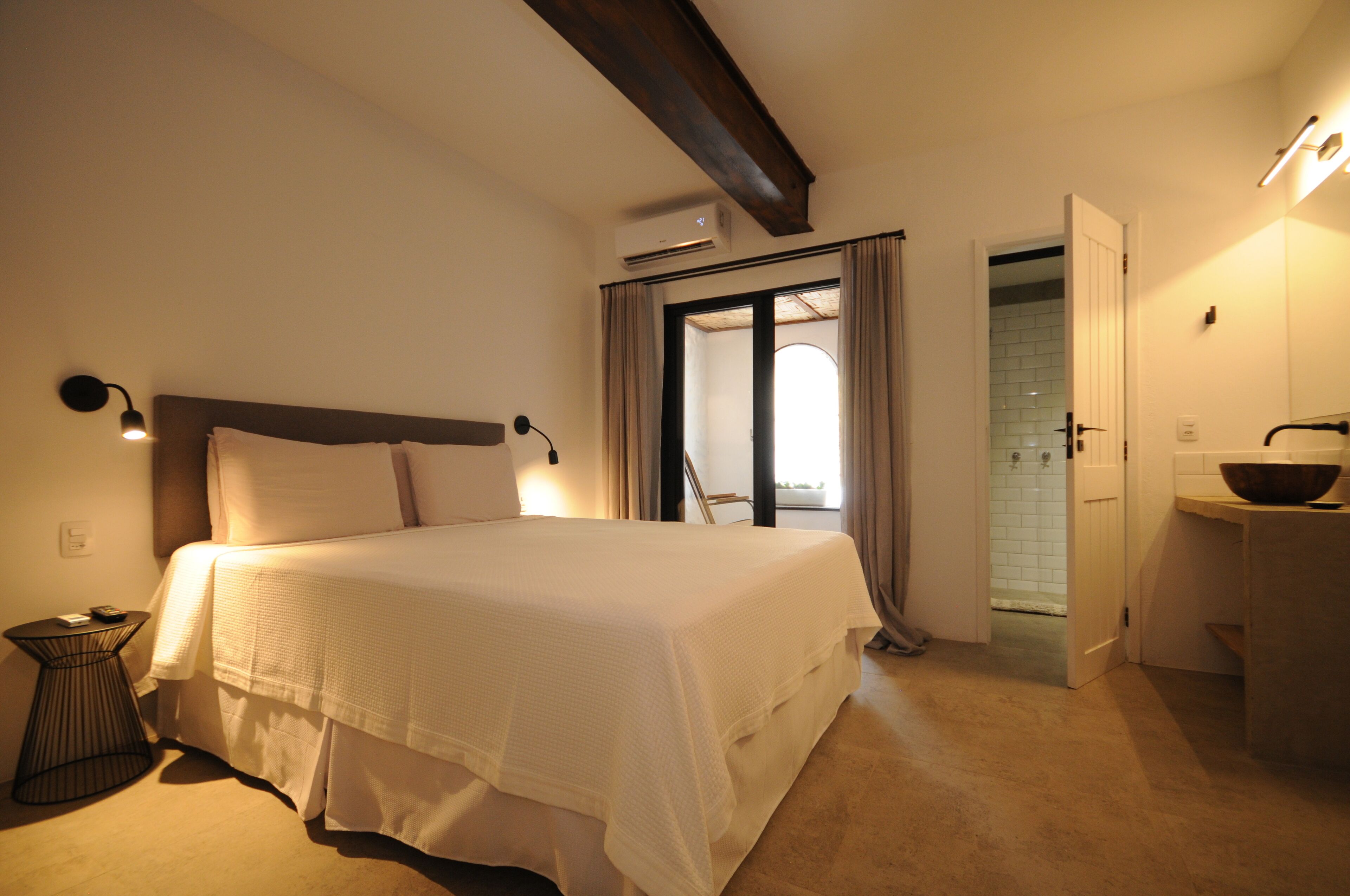 Een slaapkamer met een groot bed, een nachtkastje, een badkamer zichtbaar door een geopende deur, en een klein keukentje met een kom op het aanrecht.