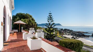 A patio with outdoor seating, a large umbrella, and a view of the ocean and mountains.