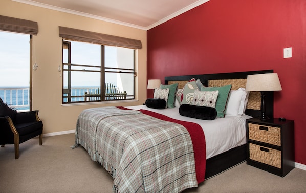 A hotel room with a large bed, a nightstand, a lamp, and a view of the ocean through a window with blinds. - Whale View Manor Guesthouse (Cape Town)