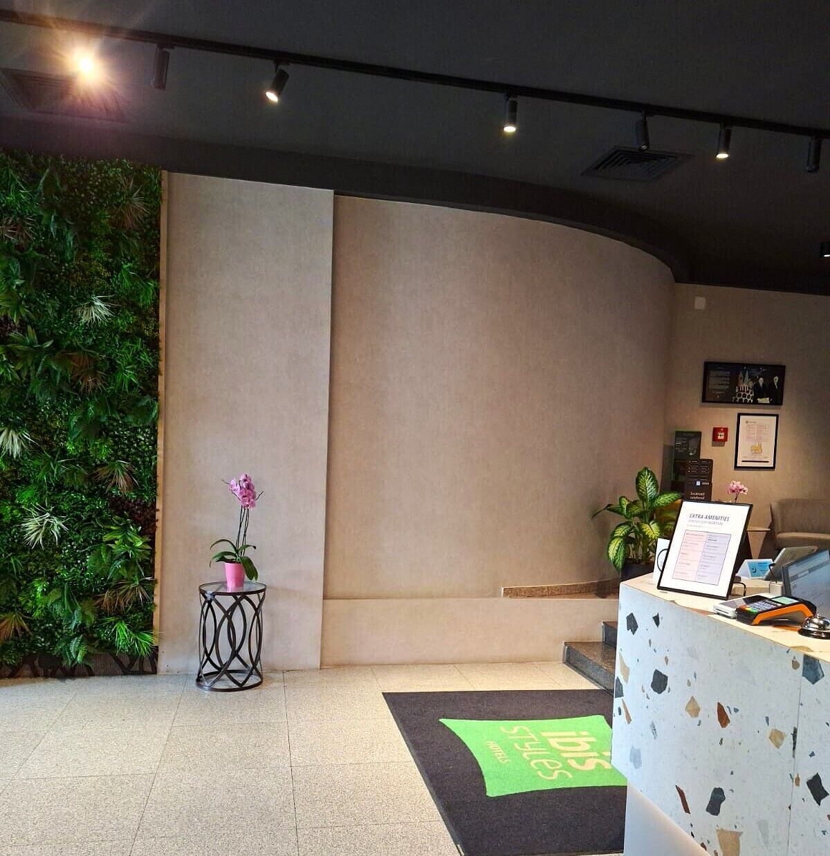 Zona de recepción con alfombra verde, mostrador de recepción, planta en maceta y pared con vegetación.