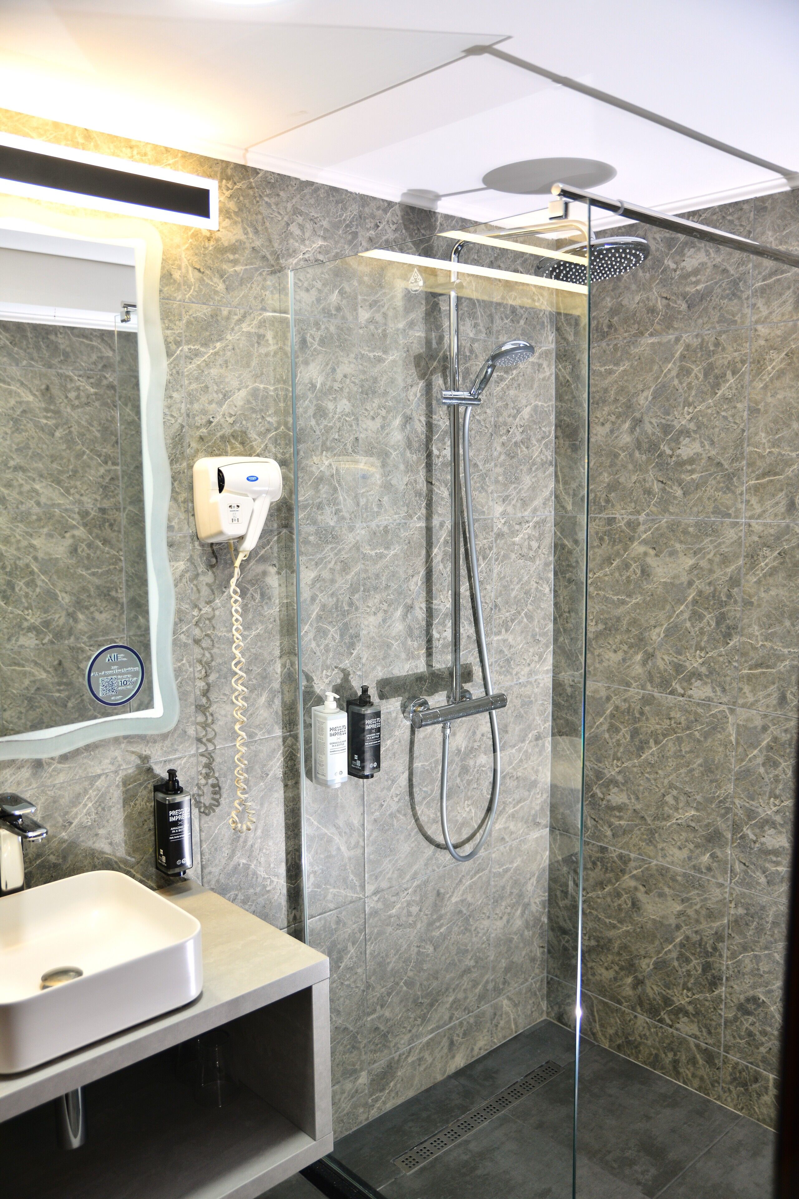Un baño moderno con ducha envuelta en vidrio, tocador con lavamanos y un secador de pelo fijado a la pared.