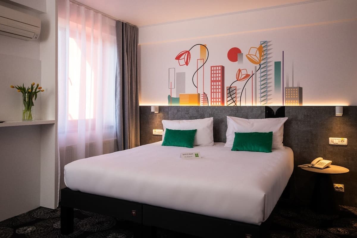 Een hotelkamer met een groot bed, twee kussens, een nachtkastje met een telefoon en een muur met abstracte kunst.