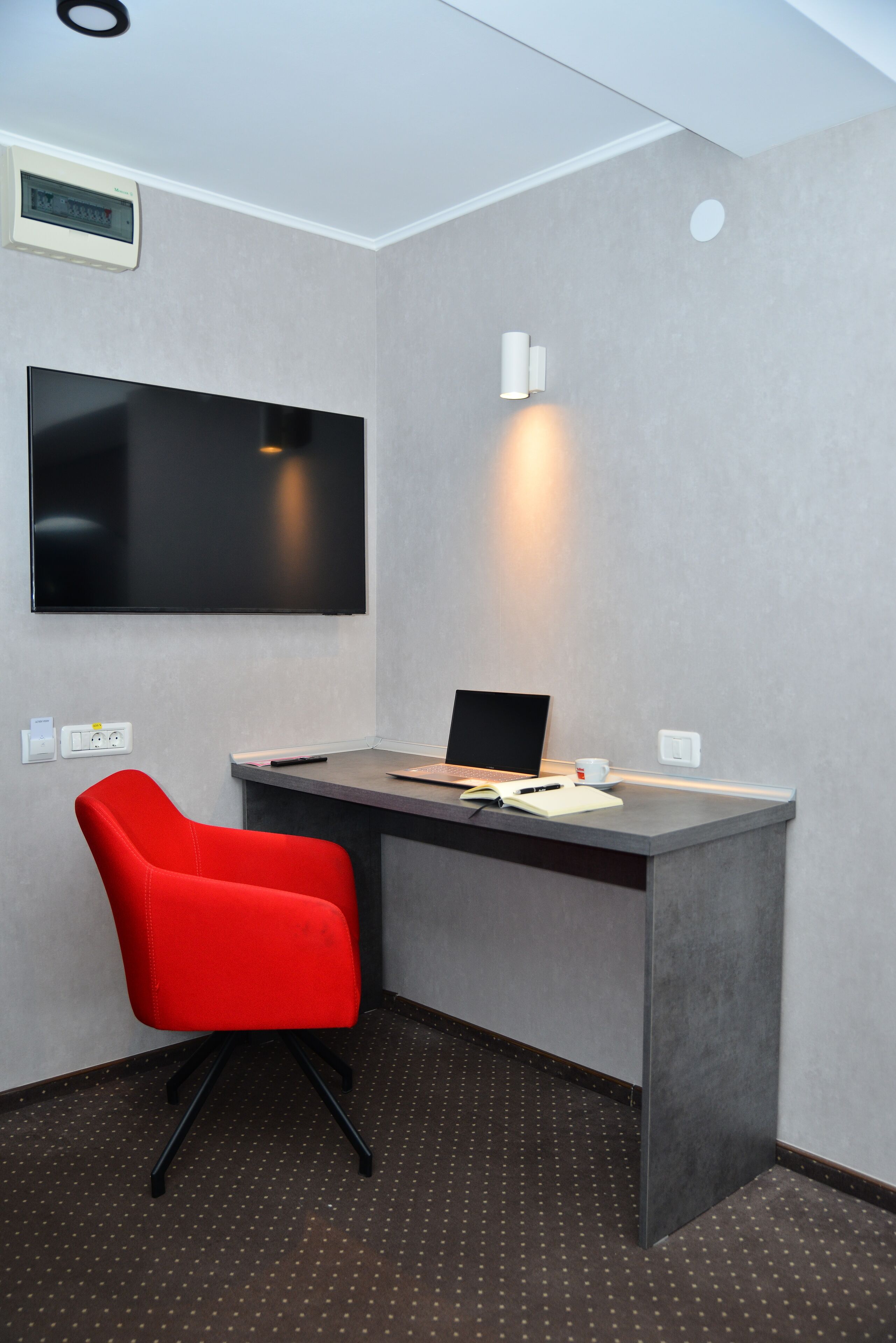 Habitación de hotel moderna con escritorio, una silla roja, un televisor de pantalla plana y una lámpara de pared.