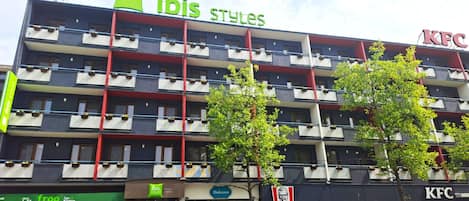 A multi-story building with 'ibis Styles' and 'KFC' signs, balconies with planters, and a 'Dolcesco' store.