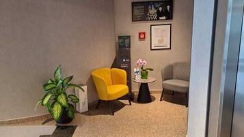 A reception area with a yellow armchair, a grey sofa, a small round table with a potted plant, and a framed picture on the wall.