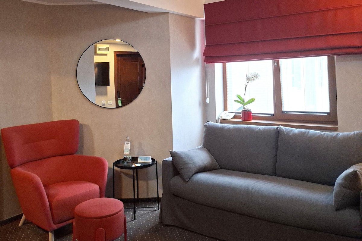 Een kamer met een grijze bank, een rode fauteuil, een klein rond tafeltje, een spiegel en een raam met een rode vouwsluier.