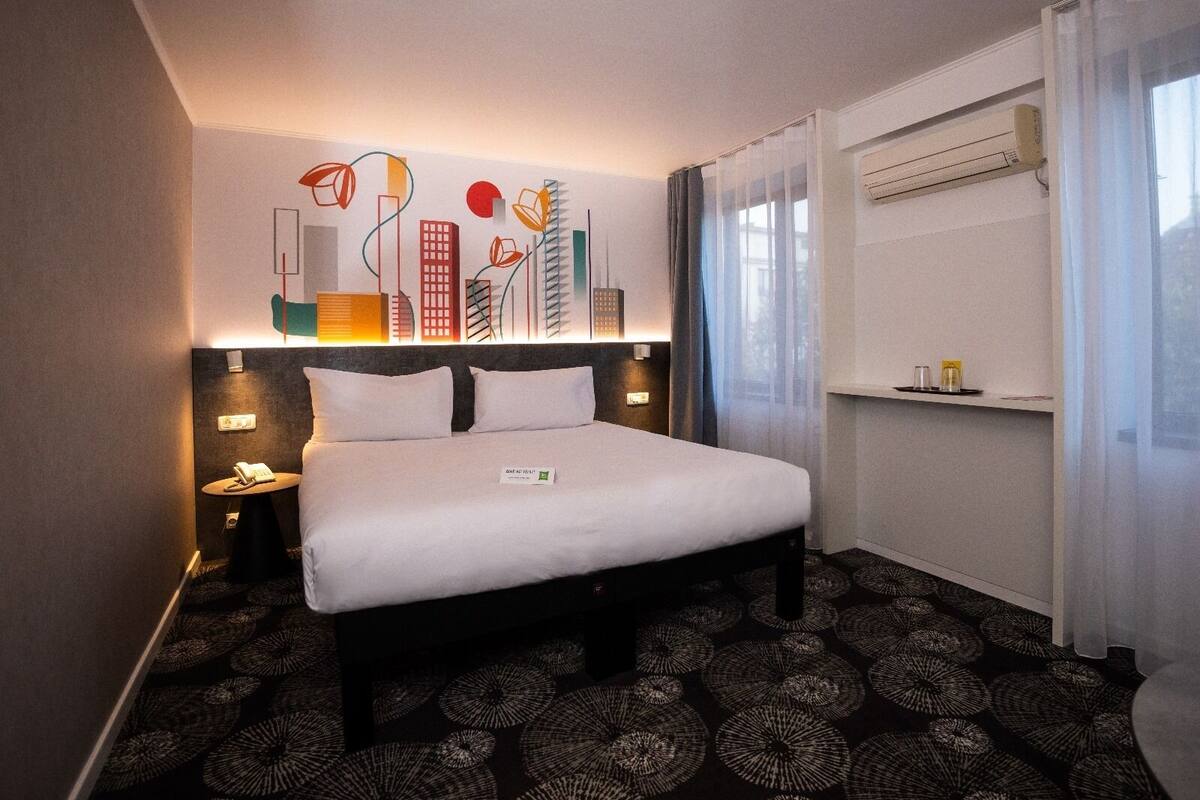 Een hotelkamer met een bed, nachtkastje, kunstwerk aan de muur en een raam met gordijnen.