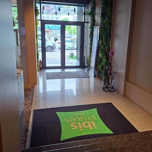 A hotel entrance with a glass door, a green mat with text, a potted plant, and a tiled floor. - Ibis Styles Pitesti Arges (Pitesti)
