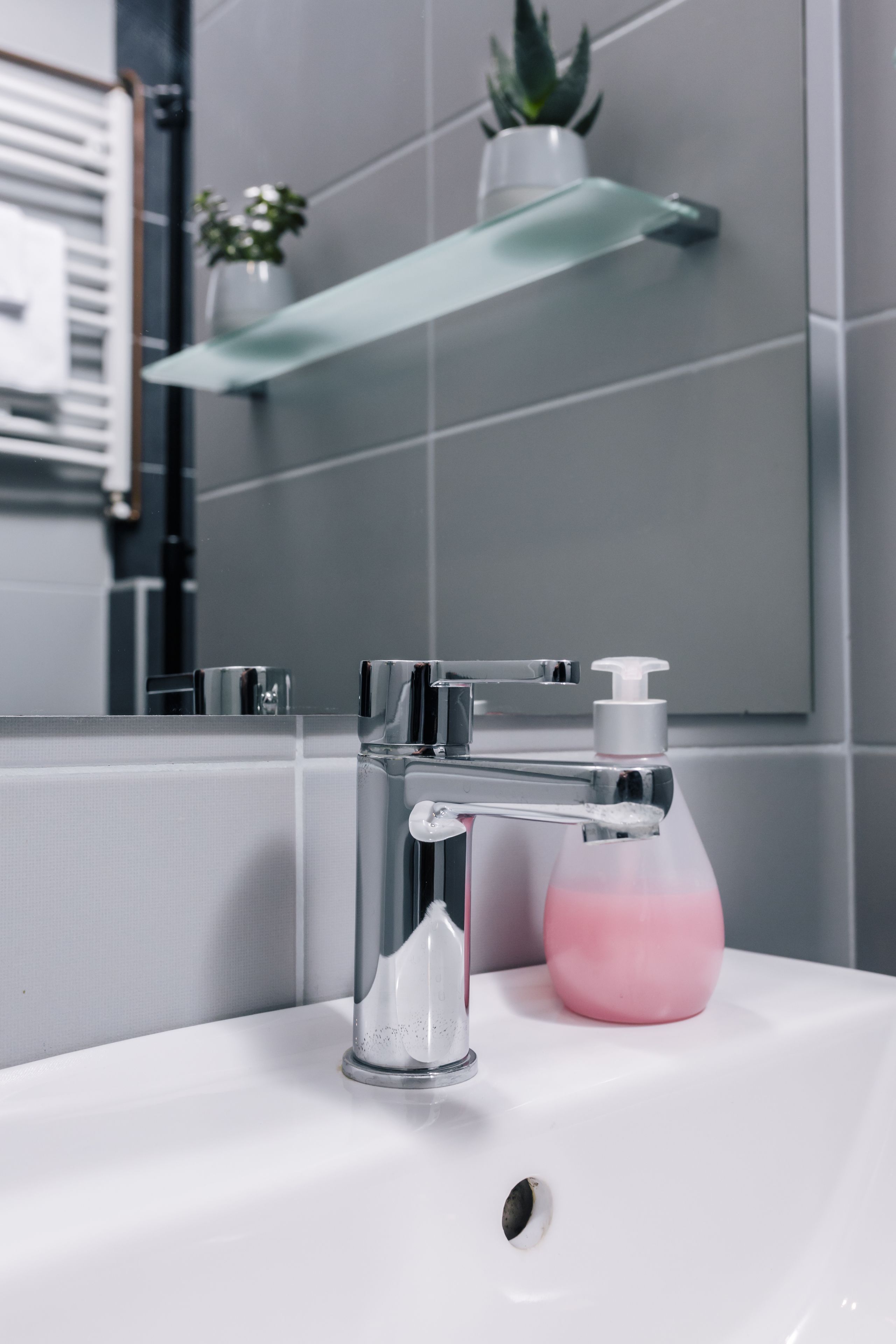 A bathroom with a chrome faucet, a white sink, a pink soap dispenser, and a glass shelf with a plant.