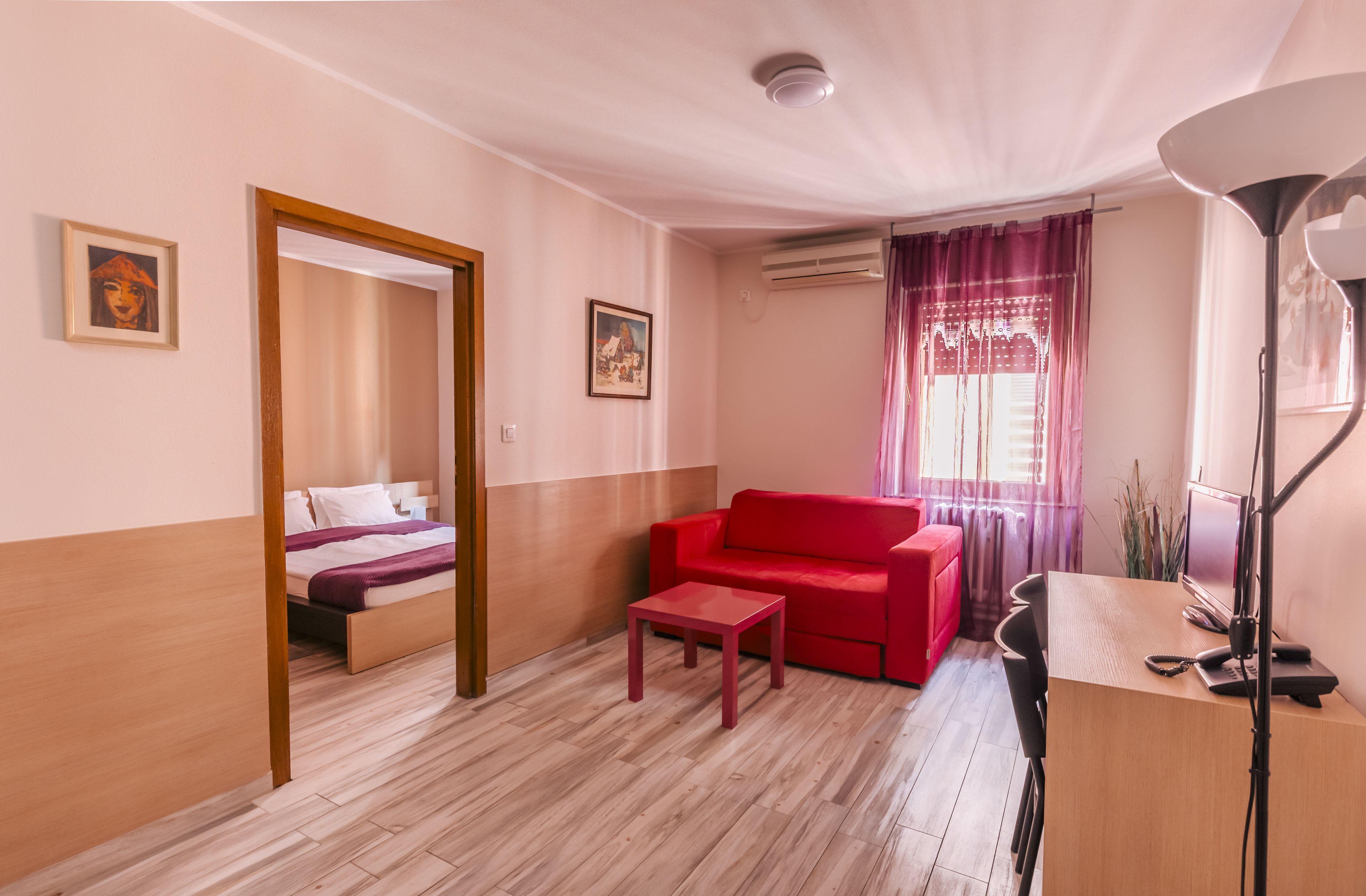 A room with a bed, a red sofa, a small table, a desk with a computer, and a lamp.