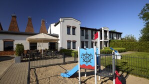 A playground with a slide and a ship-themed play structure, surrounded by a fence and a building with multiple chimneys in the background. - Kurort Stara Wędzarnia (Gdansk)