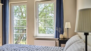 A bedroom with a patterned bedspread, two windows with blue curtains, a bedside table with a lamp, and a radiator. - Kurort Stara Wędzarnia (Gdansk)