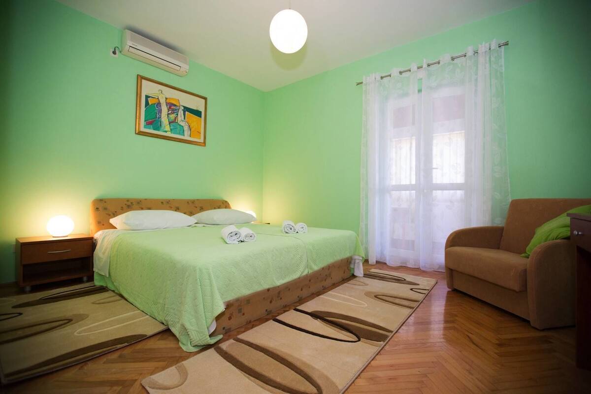 Un dormitorio con cama, mesita de noche, sofá, ventana con cortinas y un cuadro en la pared.