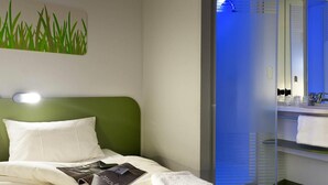 Hotellihuone, jossa on sänky, lehti, pyyhe, vihreä pääty, seinään kiinnitetty valaisin sekä kylpyhuone, jossa on peili ja pesuallas.