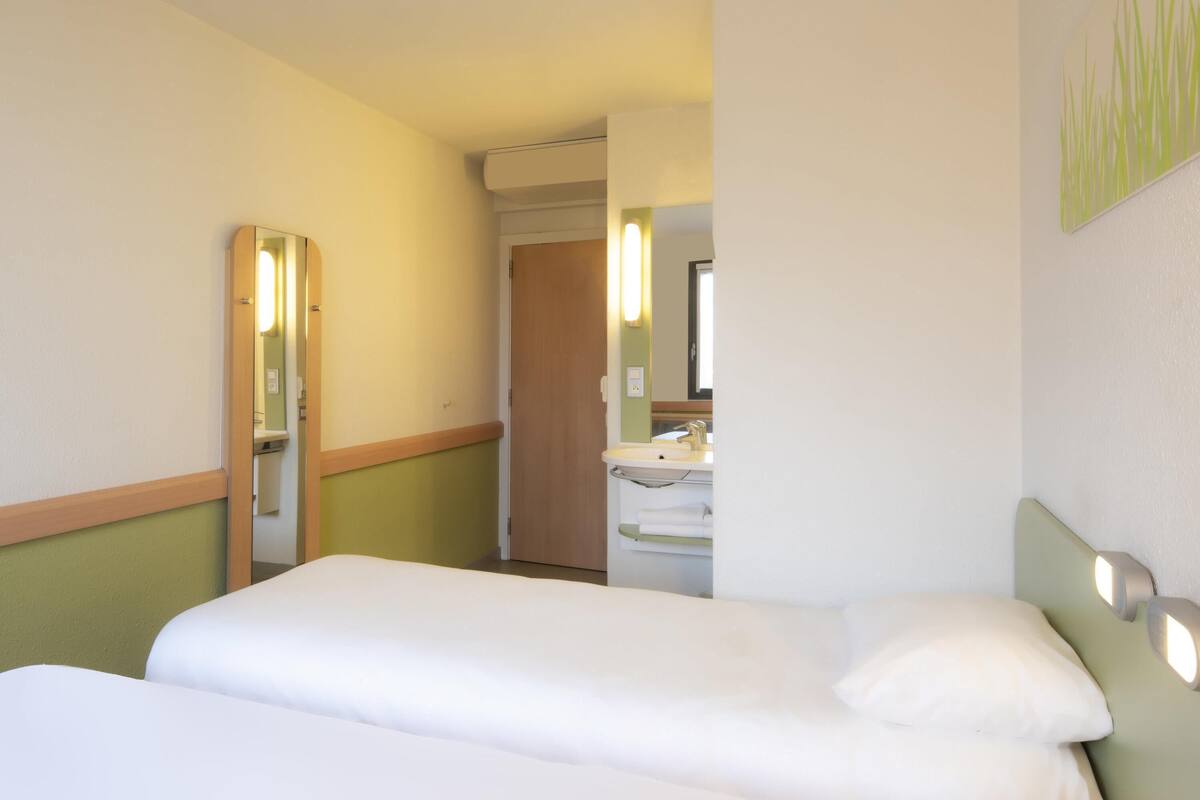 Une chambre d’hôtel comprenant un lit, une table de chevet, un miroir et une salle de bain avec un lavabo et un porte-serviettes.