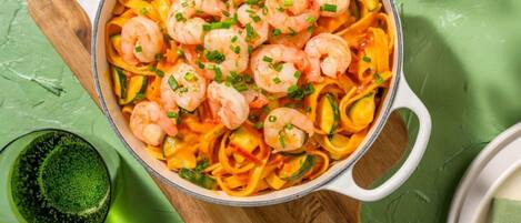 A pot of shrimp pasta with vegetables on a green surface.