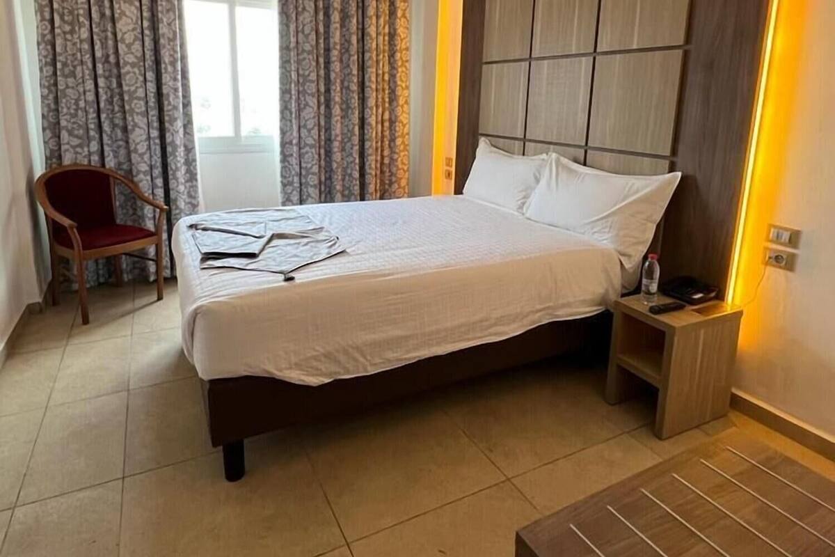 A modern hotel room with a large bed, a wooden headboard, a bedside table with a lamp, a chair, and a view of the outdoors.