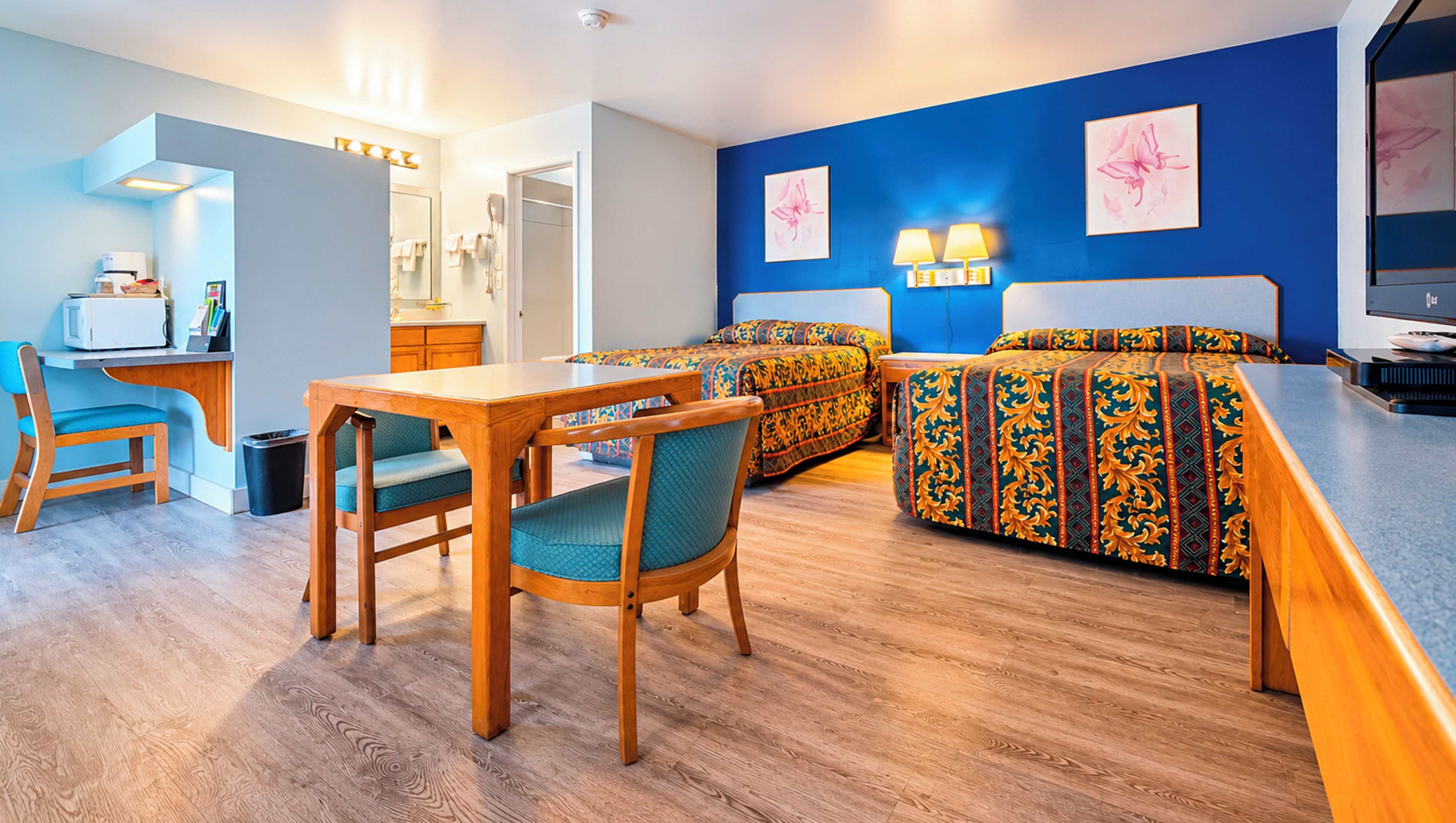 Une chambre d’hôtel avec un bureau en bois, deux lits avec une literie à motifs, un mur d’accent bleu et une petite kitchenette.