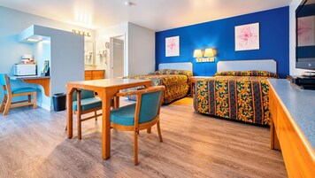 Habitación de hotel con un escritorio de madera, dos camas con ropa de cama estampada, una pared azul de acento y una pequeña cocina americana.