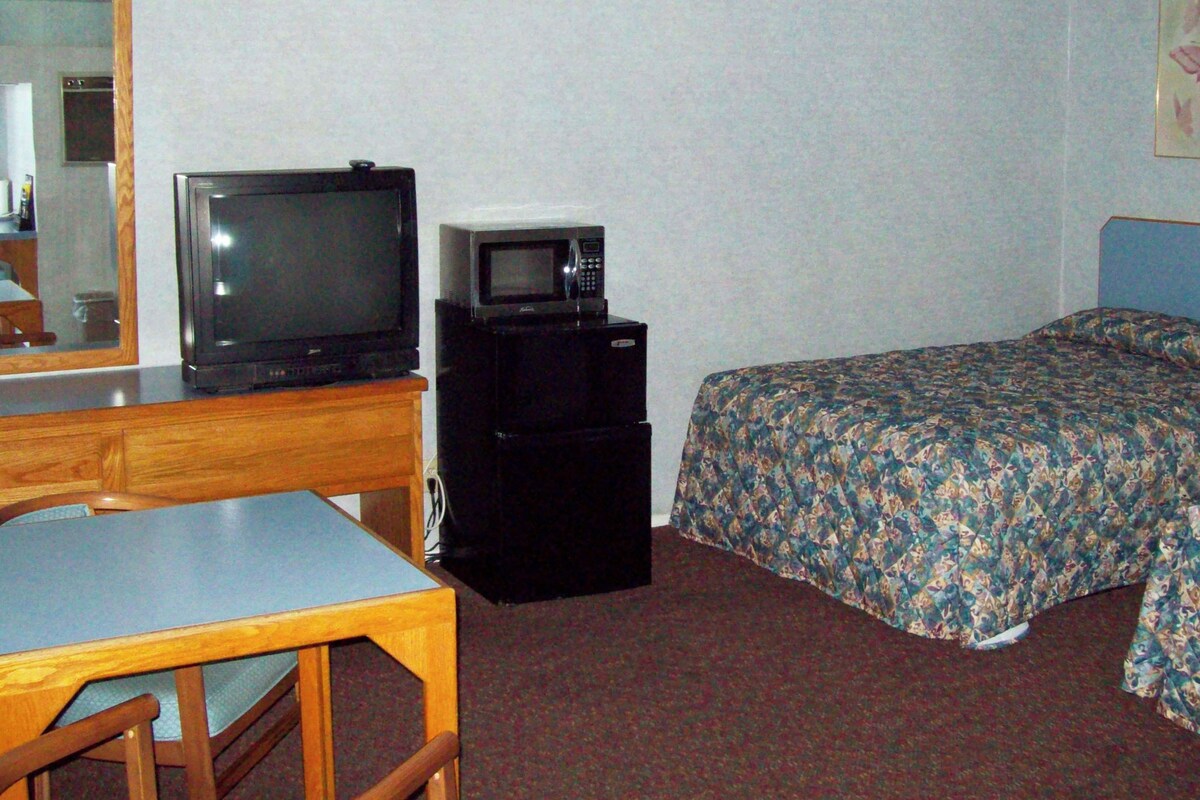 Quarto de hotel com escrivaninha de madeira, televisão, frigorífico, cama com roupa de cama floral e espelho.