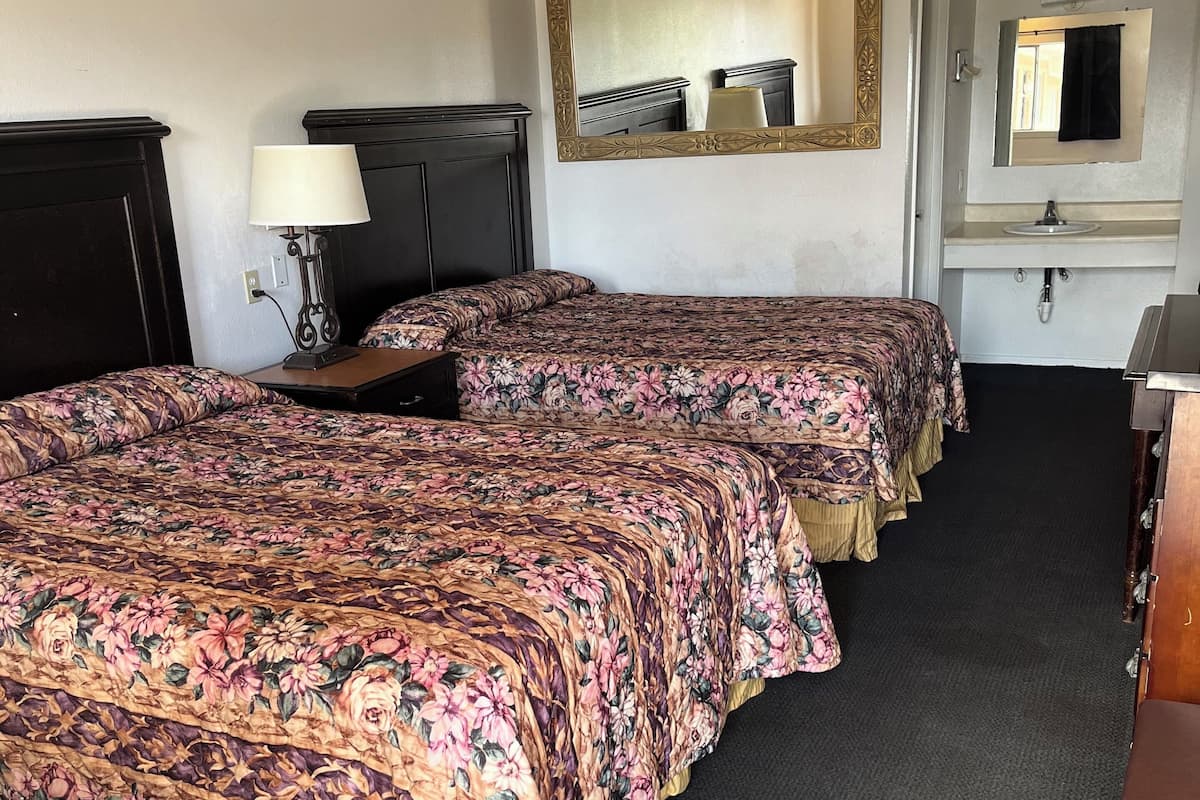 A hotel room with two beds, a nightstand with a lamp, a dresser, a mirror, and a small bathroom with a sink and mirror.