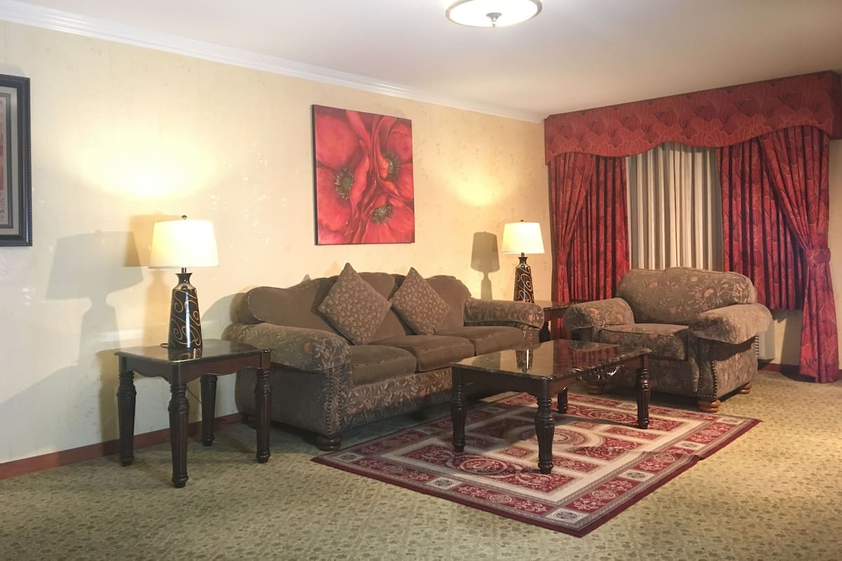 A living room with a brown sofa, two side tables with lamps, a glass coffee table, a red patterned rug, and a large red floral painting on the wall.