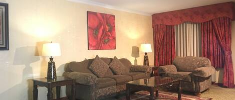 A living room with a brown sofa, two side tables with lamps, a glass coffee table, a red patterned rug, and a large red floral painting on the wall.