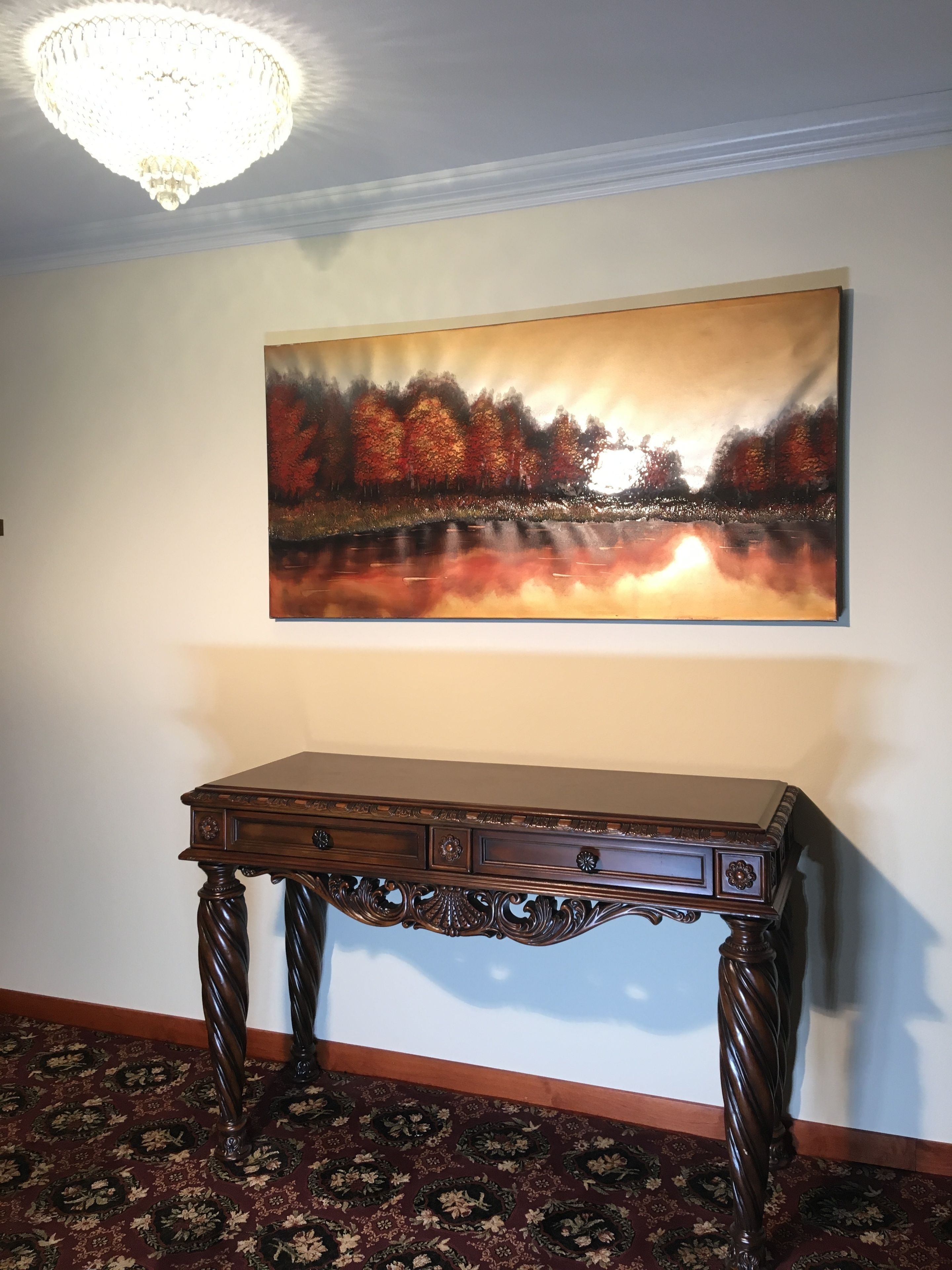 A room with a large painting of a forest scene, a wooden console table, and a patterned carpet.