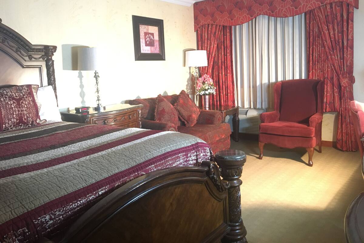 A hotel room with a large bed, a red armchair, a wooden side table, a lamp, and a window with red curtains.