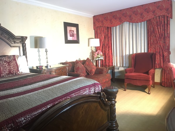 A hotel room with a large bed, a red armchair, a wooden side table, a lamp, and a window with red curtains.