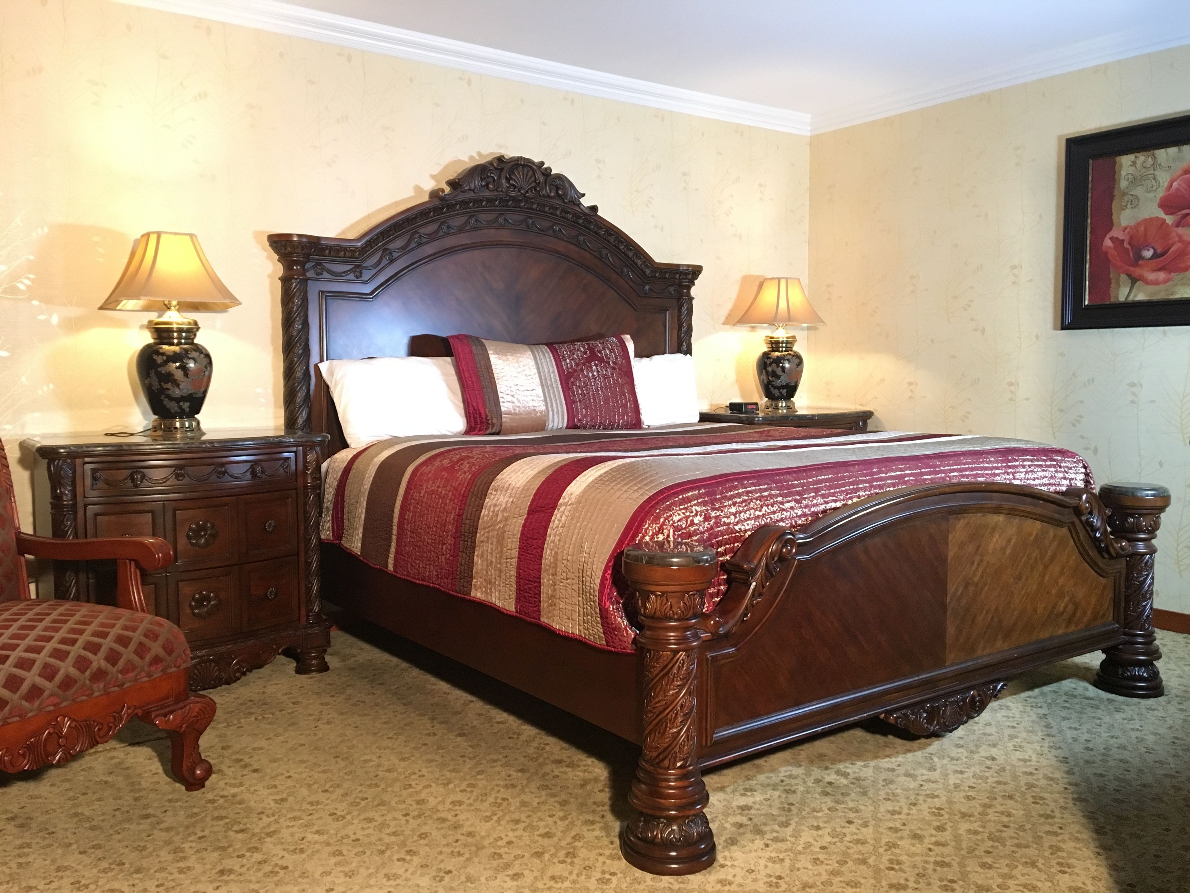 A traditional four-poster bed with a wooden headboard, flanked by two nightstands with lamps, a wooden chair, and a patterned carpet.